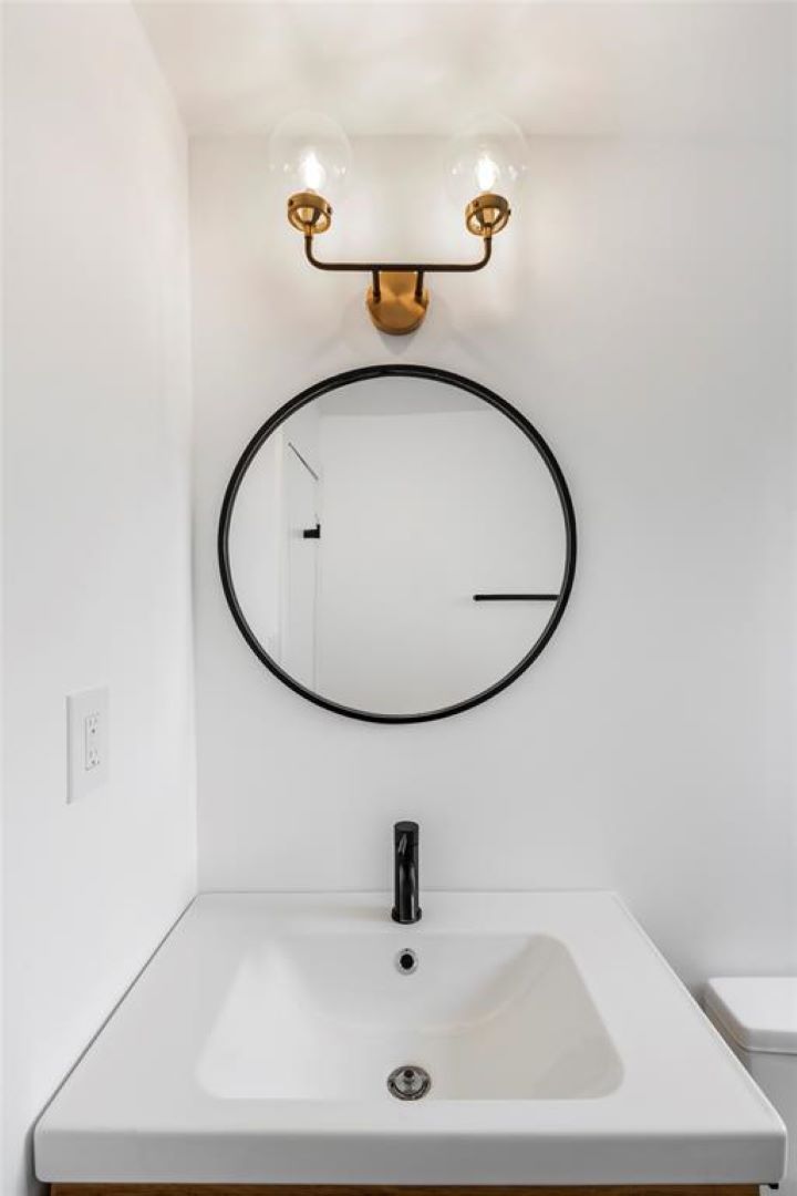 Modern powder room renovation by Houston remodeling contractor Square 1 Home Solutions, featuring a white vanity top, matte black faucet, round black-framed mirror, and a brass two-light globe sconce.